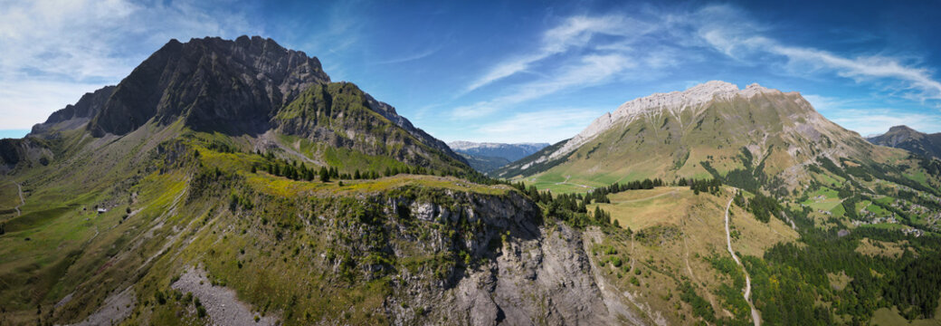 Aerial view of the majestic mountains draped in green and grey hues under a blue sky, pathways winding through the landscape, La Clusaz, Auvergne-Rhone-Alpes, France.