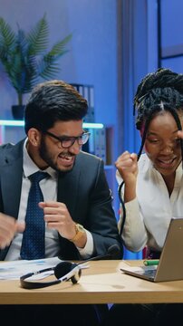 Businessman and his african american female workmate enjoying joint victory with raised hands when see fantastic information on computer monitor in workroom at late time