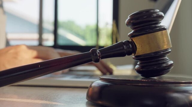 Wooden gavel rests on stand. Hand writes near gavel. Sunlight streams through window. Background shows blurred documents. Office setting suggests legal work