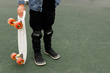 Cropped shot of a child holding a white penny board with orange wheels while standing on a dark grey rubberized playground surface, low angle view for extreme sports, childhood, and active lifestyle