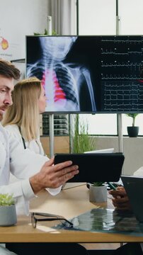 Attractive confident skilled different ages women-medics talking about patient's heart rhythm while their motivated qualified diverse male workmates reviewing information on tablet device