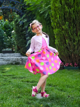 Smiling little girl in colorful polka dot dress and glasses posing in garden, happy childhood portrait outdoors,