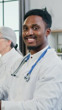 Portrait of likable positive professional modern african male medical worker which looking into camera with friendly face expression while another workmates holding discussion in medical room