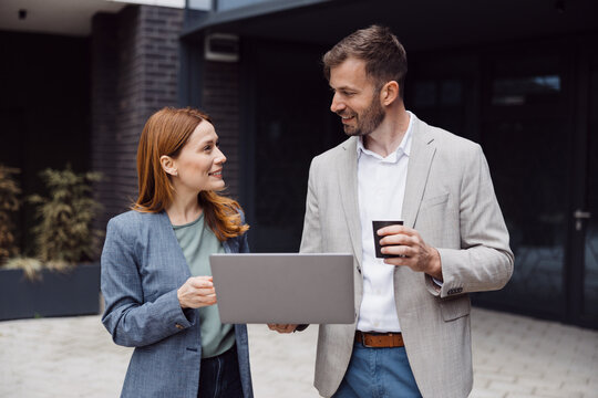 Two young professional colleagues standing or sitting outside a modern office building, working on their laptops and holding takeaway coffees