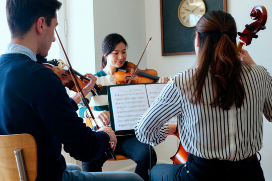 Young adult musicians rehearsing string ensemble with violins and cello while reading sheet music in classroom