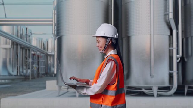 Side View of Asian Female Engineer Use Computer While Walking at Industrial Chemical Plant with Row of Stainless Steel Storage Silos