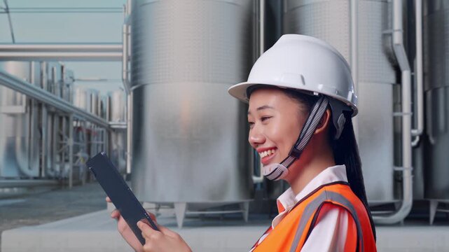 Side View of Asian Female Engineer Use Tablet While Walking at Industrial Chemical Plant with Row of Stainless Steel Storage Silos