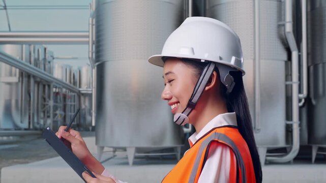 Side View of Asian Female Engineer Taking Note on Tablet While Walking at Industrial Chemical Plant with Row of Stainless Steel Storage Silos