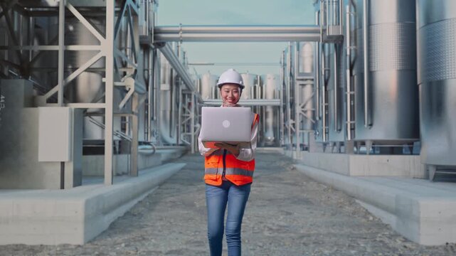 Front View of Asian Female Engineer Use Computer While Walking at Industrial Chemical Plant with Row of Stainless Steel Storage Silos