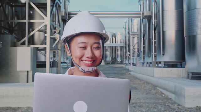 Front View of Asian Female Engineer Use Computer While Walking at Industrial Chemical Plant with Row of Stainless Steel Storage Silos