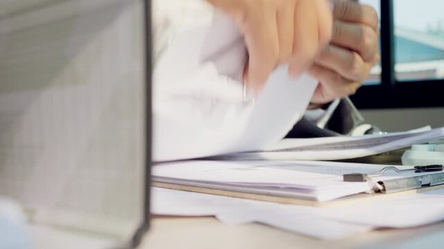 Hand organizes documents near a laptop. Fingers flip papers with focused precision. Office work shown in close-up detail