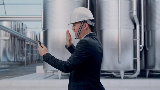 Side View of Asian Businessman Use Smartphone, Video Call While Walking at Industrial Chemical Plant with Row of Stainless Steel Storage Silos