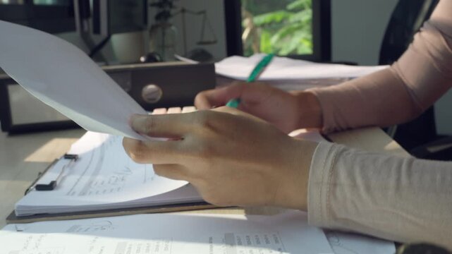 Woman calculates data with a handheld calculator. She writes notes on documents with a green pen. Natural light illuminates her workspace