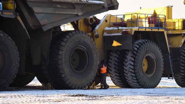 Worker in High-Visibility Vest Inspecting Mining Dump Truck Wheels in Winter. Maintenance Engineer Next to Heavy Industrial Vehicle at a Mining Site