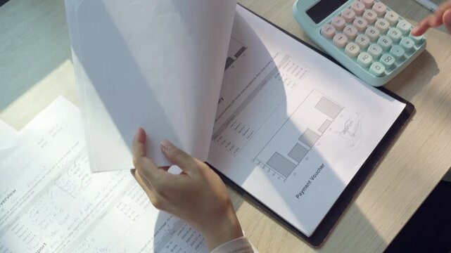 Hands flip papers over a desk. Sunlight glints off charts and data sheets. Calculator rests nearby, unused. Fingers trace graphs with a green marker. Office work feels precise and focused