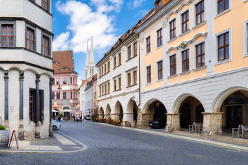 Fototapeta premium View from the Untermarkt in Görlitz toward the spires of St. Peter and Paul church between historic facades, cobblestone pavement and blue sky with clouds.
