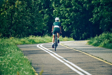 Obraz premium A woman cyclist in green gear rides away along a path surrounded by flowering trees, captured from behind at a wider distance. The image conveys freedom, exploration, and the beauty of summer.