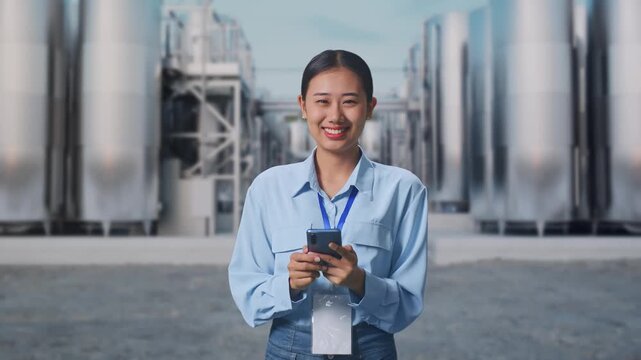 Asian Female With Her Smarphone at Chemical Plant with Row of Stainless Steel Storage Silos, She Is Looking At The Camera With A Smile