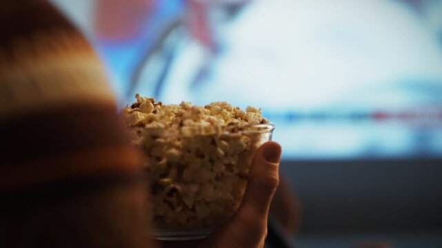 A person enjoys a bowl of popcorn while watching an engaging movie on a large screen. This relaxing moment occurs in a cozy living room setting, perfect for entertainment and leisure