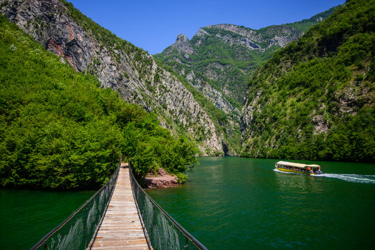 A rustic wooden suspension bridge spans the emerald Koman Lake, with a yellow tour boat traversing the water amidst steep, verdant mountains in Albania