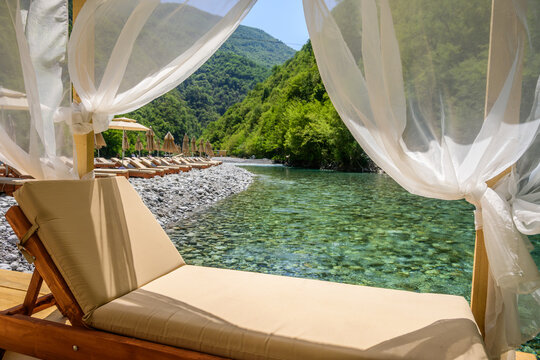 A serene riverside setting with sun loungers and straw umbrellas lining a rocky bank, overlooking the transparent turquoise Shala River and lush green mountains in Albania