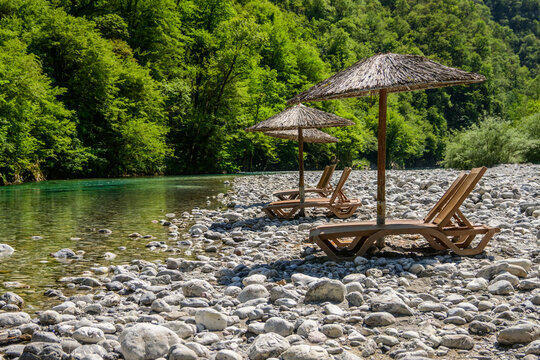 Rustic straw umbrellas and wooden loungers are set on a rocky riverbank beside the transparent turquoise Shala River, with dense green mountains as a backdrop in Albania