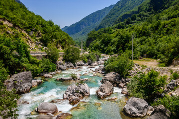 A vibrant Valbona Valley river with stunning turquoise water cascades over rocks, surrounded by densely forested mountains under a clear blue sky, evoking wilderness in Albania. © Danica Chang