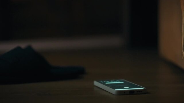 A man's hand reaches for a phone that lights up while it's on the floor next to his bed. Smartphone alarm in the morning. Fingers turn off the alarm