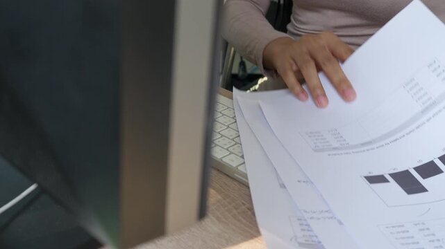 Hand holds papers near keyboard. Person works at desk with monitor. Fingers press keys, focus on task. Office setting, no animals or plants