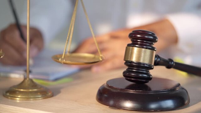 Wooden gavel rests beside golden scales. Hand adjusts scales in soft focus background. Legal documents lie beneath the gavel and scales