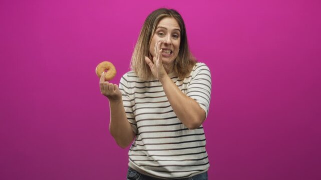 Woman holding donut and touching cheek with wry face in studio, striped shirt and visible hand gesture against pink backdrop; temptation curiosity.