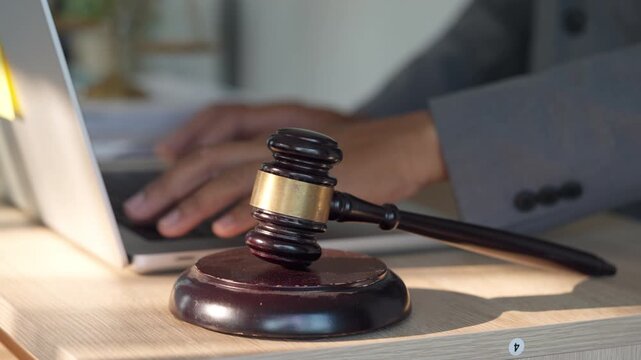 Wooden gavel rests beside open laptop. Hands type on keyboard, showing legal work. Gavel symbolizes judgment, law, and authority. Office desk setup suggests modern legal practice