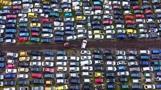 A junkyard filled with many parked cars of various colors and conditions sits under daylight. Rows of vehicles are closely packed along dirt paths.