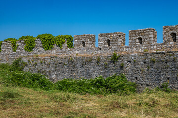 An expansive view of the robust stone walls and battlements of Rozafa Castle, partly covered in lush green ivy, standing under a clear blue sky in Shkoder, Albania. © Danica Chang