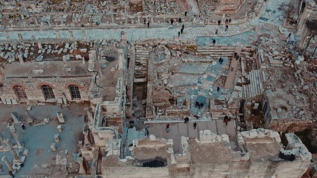 Vertical top-down drone shot of the marble-paved streets and surrounding ruins in Ephesus, Izmir, Turkey. Visitors walk along the ancient pathways, revealing the layout of Roman urban planning and arc