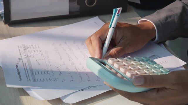 Hands calculate figures on papers with a calculator. Documents lie stacked on a wooden desk surface. One hand writes, another holds a pen. Hands adjust papers while working on a calculator