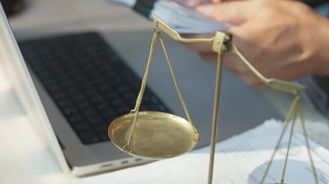 Balanced scales rest near a laptop. Hand types on keyboard beside the scales. Work surface holds documents and tech. Symbolic legal balance in modern setting. Focus on scales, hands, and digital tools