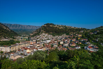 Fototapeta premium A broad, vibrant panorama of Berat, Albania, captured on a sunny day, featuring the city, the Osum River, and the forested castle hill against distant mountains.