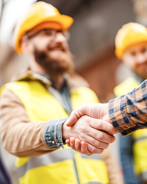 Two construction workers wearing yellow helmets and safety vests shake hands, symbolizing agreement or partnership on a worksite.