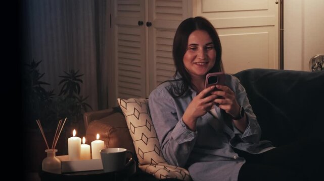 Happy woman using smartphone at home in cozy living room. Smiling young woman subscribing to new social media, shopping online, ordering products online using apps, watching series, chatting with frie