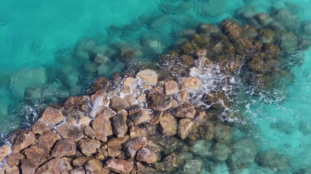 Aerial drone view of waves washing over large rocks in crystal clear turquoise water along the coast