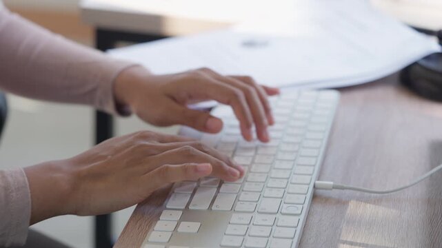 Hands type on white keyboard at wooden desk. Fingers press keys with focused rhythm. Documents lie beside keyboard, slightly blurred. Soft sunlight hits desk surface gently