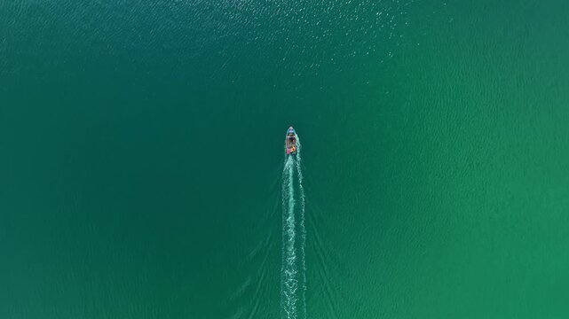 A boat travels through blue water under direct sunlight. The scene shows ripples behind the boat as it moves. Surrounding water is clear and calm with no visible land nearby.