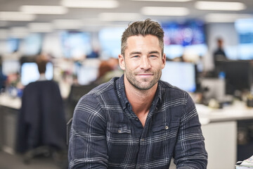 Fototapeta premium A man in a plaid shirt smiles confidently in a modern office environment with blurred computers and coworkers in the background.
