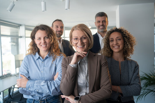 Happy diverse business team standing in the office. Five cheerful confident successful people in smart casual suits smiling and posing for a group photo all together