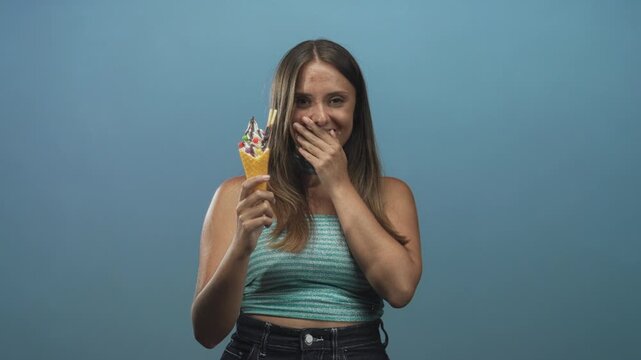 Young woman holding ice cream cone and covering mouth while laughing in studio wearing turquoise crop top and jeans; joy youthfulness.