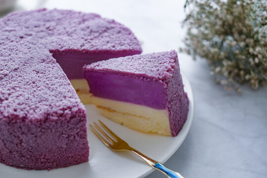 A delicious slice of layered taro cheesecake on a white plate with a gold fork, alongside the rest of the cake.