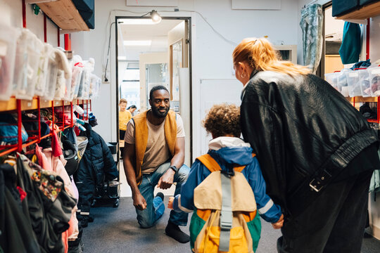 Male teacher kneeling down to greet boy and his mother in kindergarten hallway