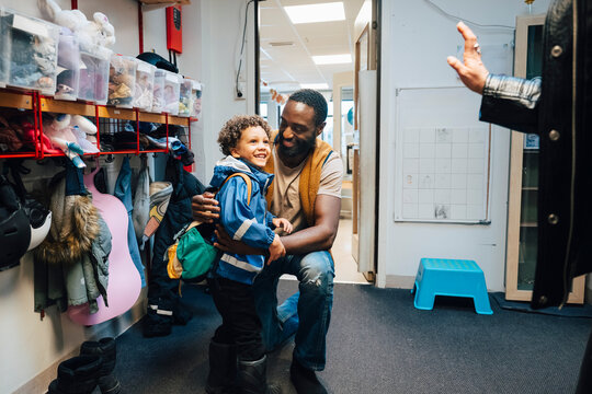 Smiling teacher embracing boy looking at his mother waving goodbye during first day of school