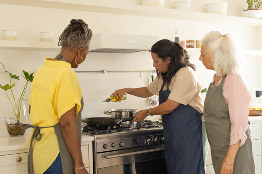 Diverse women with senior in aprons pouring oil into frying pan on gas range at home